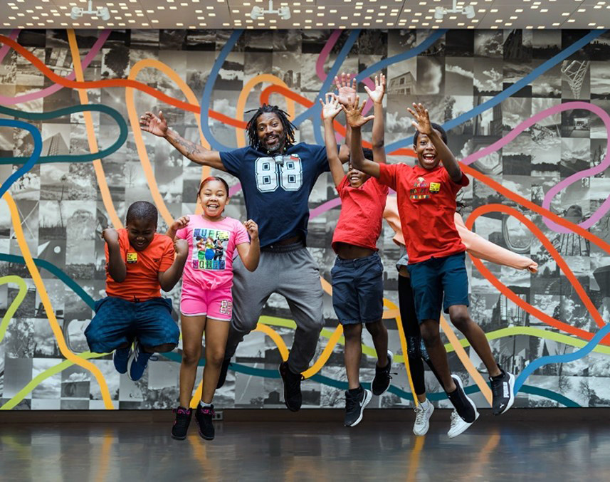 Image 1: A group of children and one adult happily jumping in front of a colored wall in the Denver Art Museum.